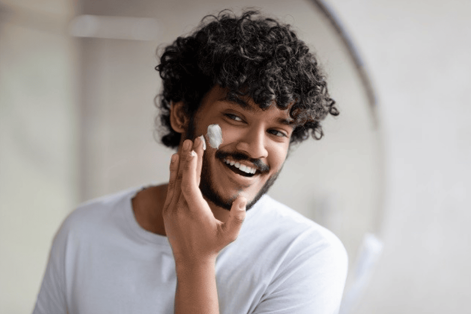 Pria berambut keriting tersenyum sambil mengaplikasikan pelembap atau skincare di wajah, ilustrasi basic skincare routine untuk cowok pemula.