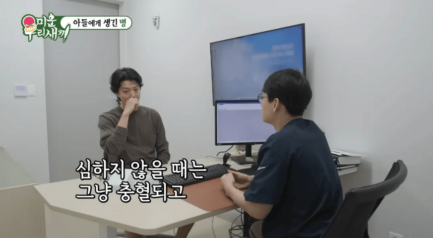 Lee Dong-gun dengan ekspresi serius berbicara dengan seorang dokter di dalam ruangan, di mana layar monitor menampilkan informasi medis.