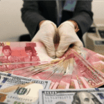 Tangan memegang Dolar AS pecahan $100 di atas tumpukan Rupiah Rp100.000, transaksi valuta asing.