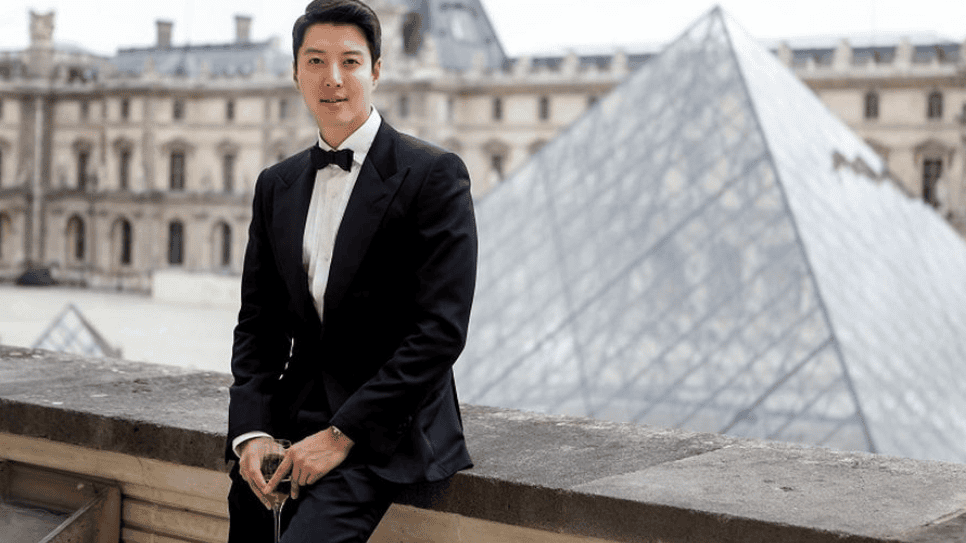 Aktor Lee Dong-gun mengenakan setelan jas hitam dan dasi kupu-kupu, berpose dengan latar belakang Piramida Louvre di Paris.