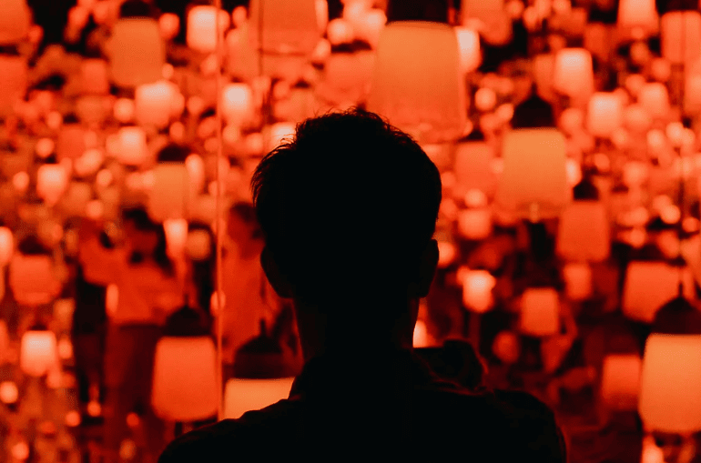 Siluet pengunjung di tengah instalasi lampu atau lampion gantung berwarna oranye, nuansa Festival Lampion.