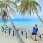 Pulau Banyak Aceh: Hidden Paradise Indonesia di Ujung Sumatera yang Mulai Dilirik Dunia Pria berwisata di pantai tropis, duduk di jembatan kayu miring dikelilingi pohon kelapa dan laut biru.