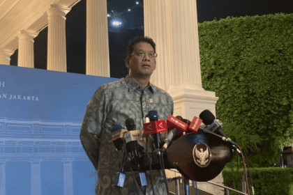Menteri Keuangan Purbaya Yudhi Sadewa mengenakan kemeja batik biru berdiri di podium dengan banyak mikrofon dan lambang Garuda Pancasila untuk memberikan keterangan pers